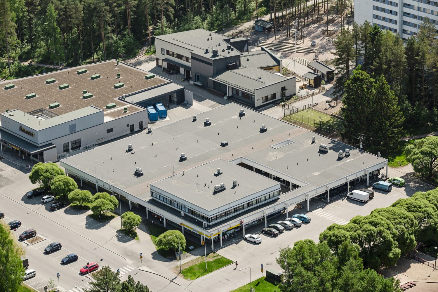 Korkealta otettu ilmakuva liikekeskuksesta, sen parkkipaikasta ja ympäröivästä metsäisestä asuina...