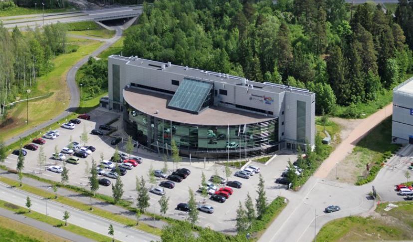 Ilmakuva modernista, kaarevasta toimistorakennuksesta ja sen pysäköintialueesta.