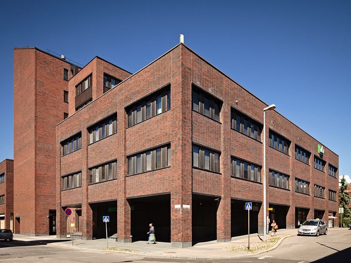 Monikerroksinen punatiilinen rakennus kadunkulmassa aurinkoisena päivänä.