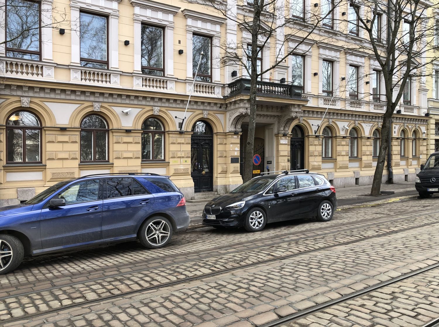 Pysäköityjä autoja vanhan rakennuksen edustalla mukulakivikadulla, jossa on raitiovaunukiskot.