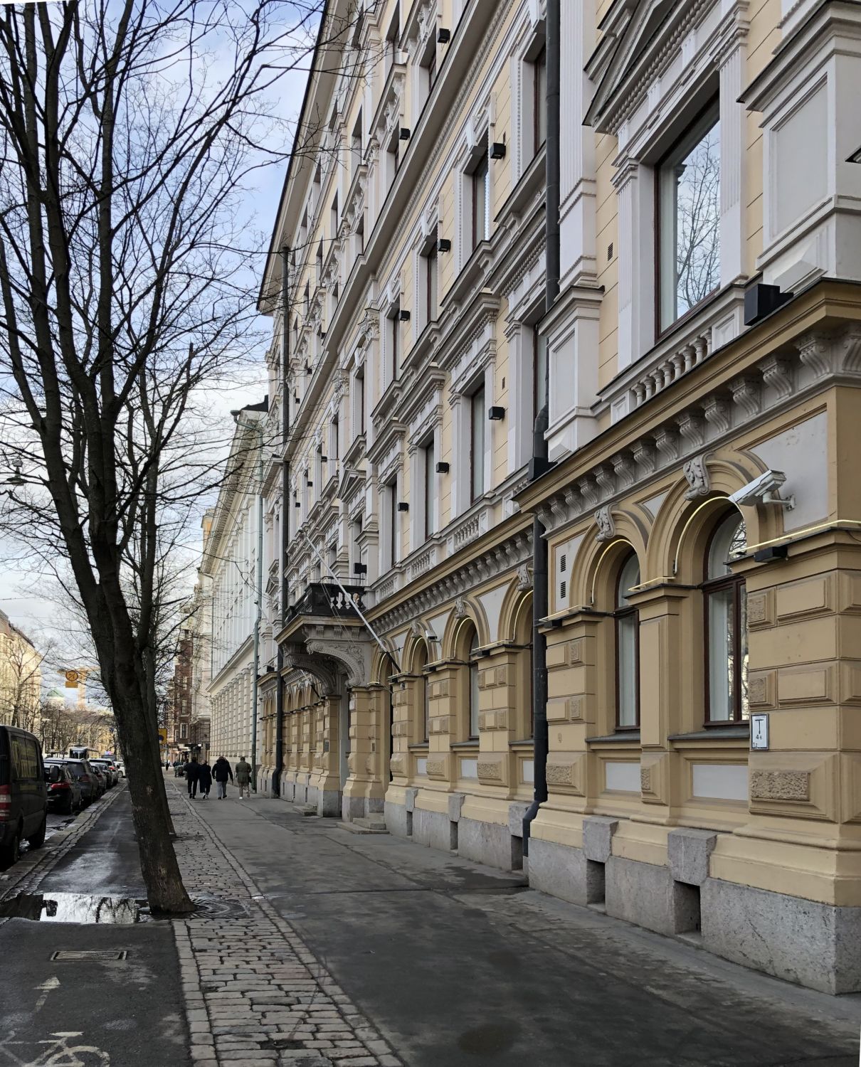 Katunäkymä, jossa on koristeellinen vaaleankeltainen rakennus ja paljas puu jalkakäytävän reunassa.