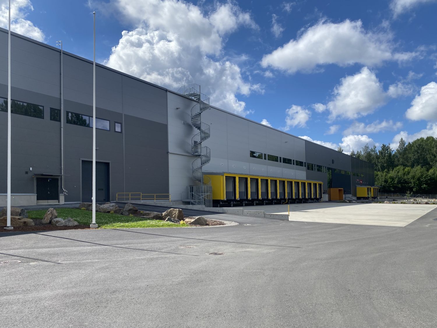 Moderni harmaa logistiikkakeskus keltaisilla lastauslaitureilla ja ulkopuolisilla kierreportailla...