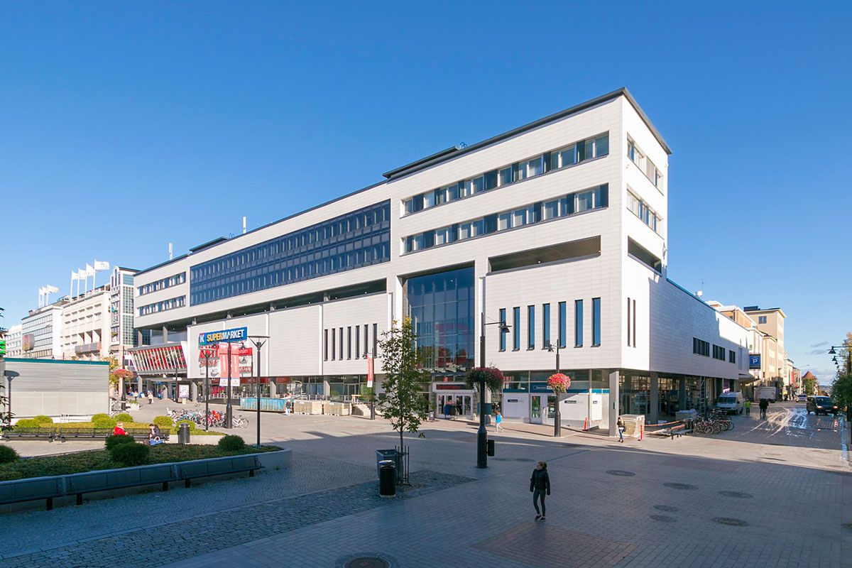 Moderni valkoinen liikerakennus ja kivetty aukio aurinkoisena päivänä.