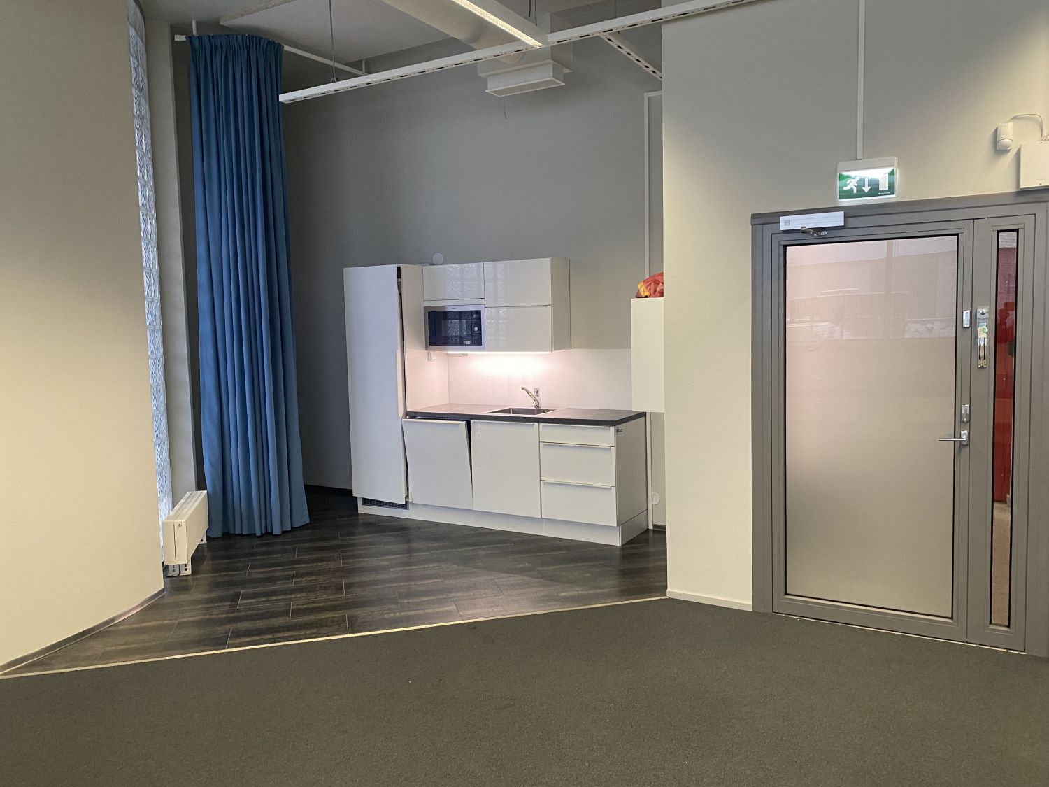 Valkoinen pienkeittiö toimistotilassa sinisen verhon ja harmaan oven välissä.