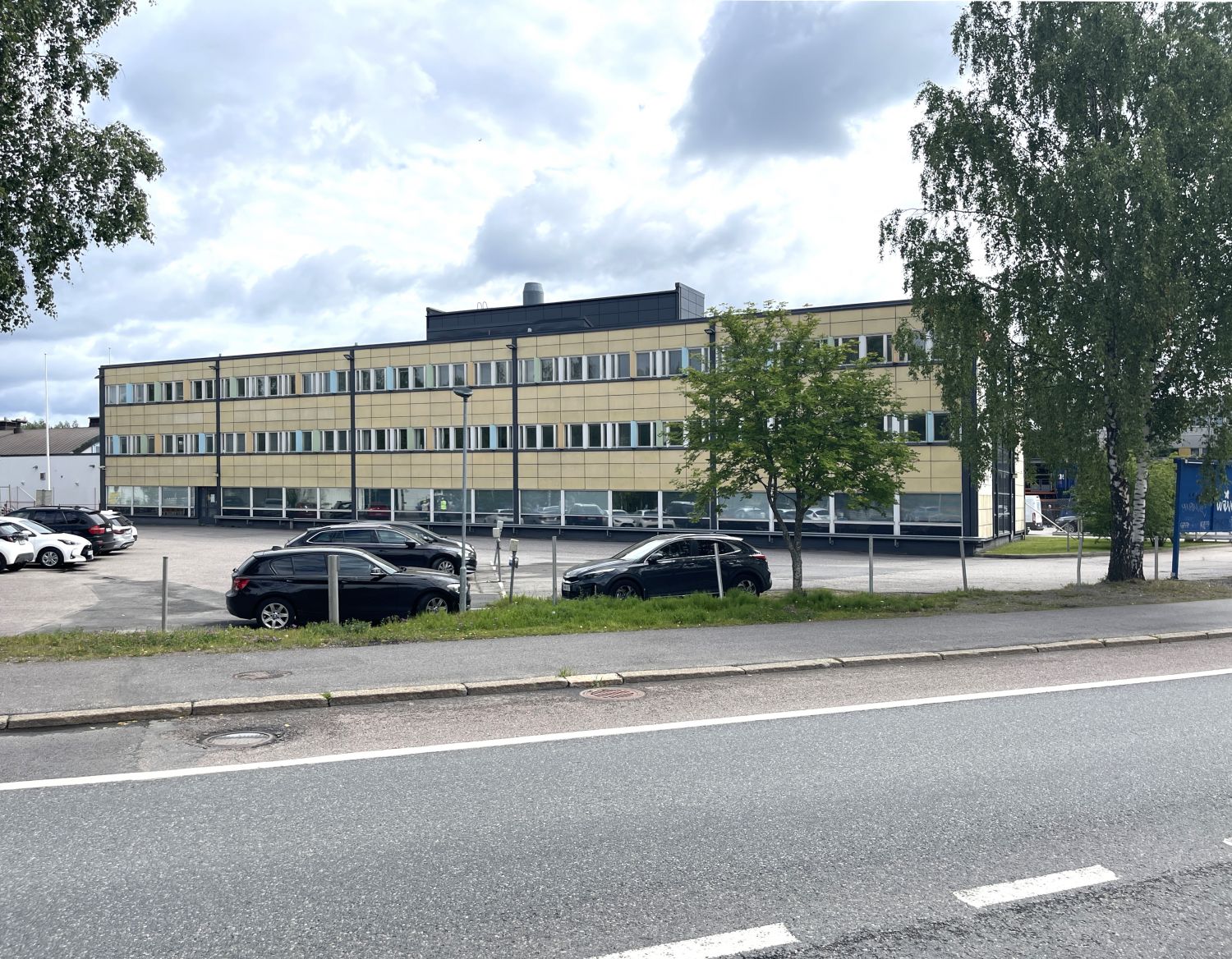 Kolmikerroksinen keltainen rakennus, jonka edustalla on parkkipaikka ja etualalla asfaltoitu tie.