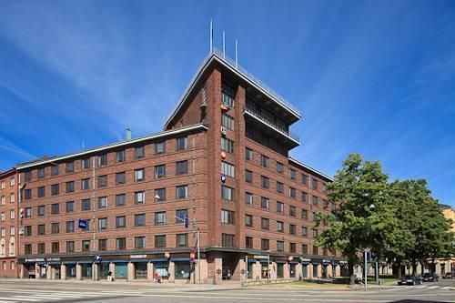 Monikerroksinen punatiilinen rakennus kadun kulmassa aurinkoisena päivänä.