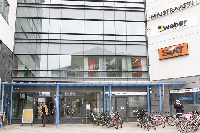 Toimistorakennuksen sisäänkäynti, jonka edessä on polkupyöräparkki ja yritysten kylttejä.