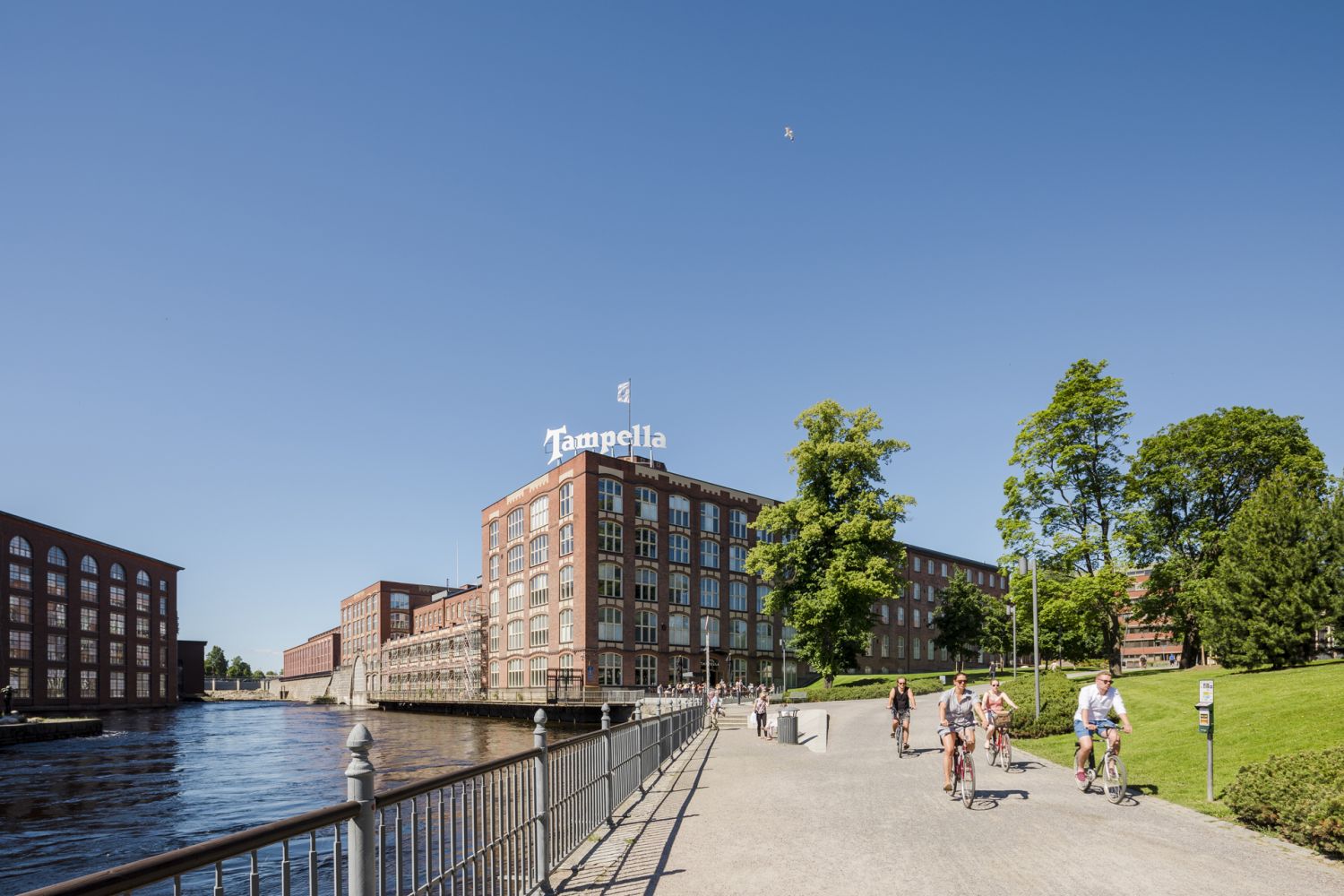 Pyöräilijöitä kulkemassa rantaa pitkin vanhojen punatiilisten tehdasrakennusten ohi aurinkoisena ...