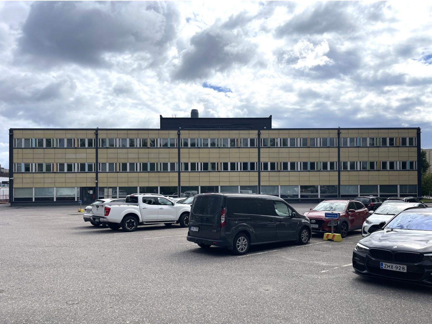 Kolmikerroksinen keltainen toimistorakennus ja sen edustalla oleva parkkipaikka autoineen pilvise...
