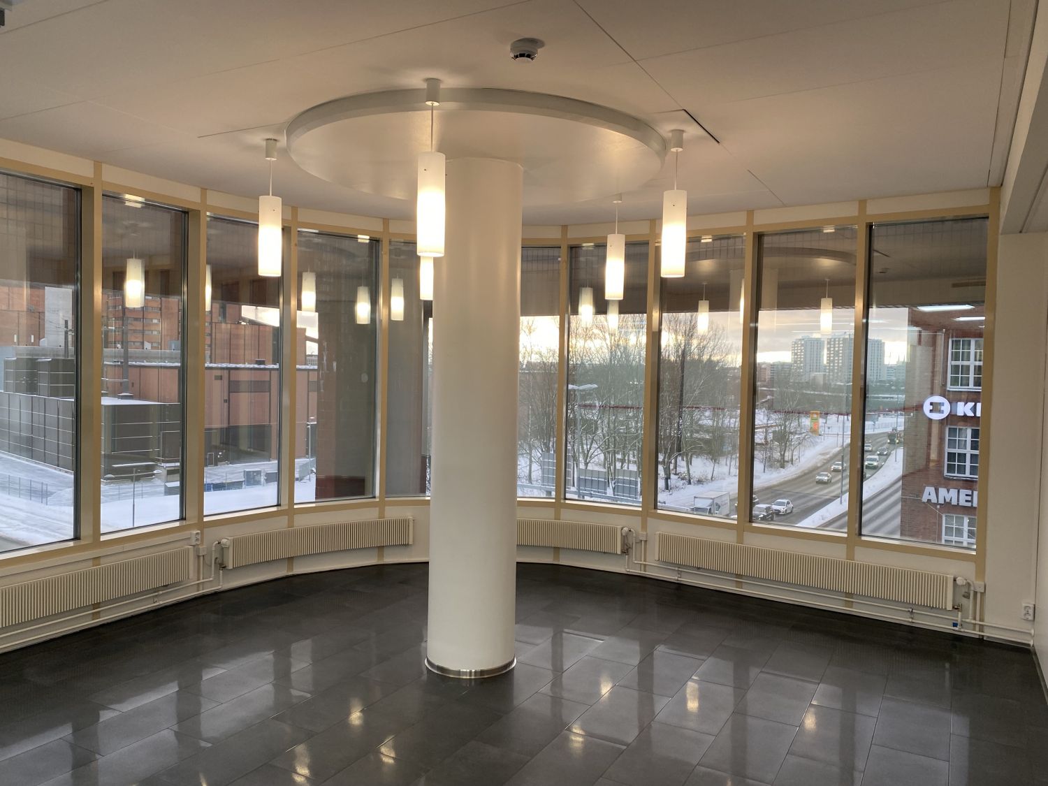 Valoisa pyöreä huone, jossa on suuret ikkunat, valkoinen keskuspylväs ja riippuvalaisimia.