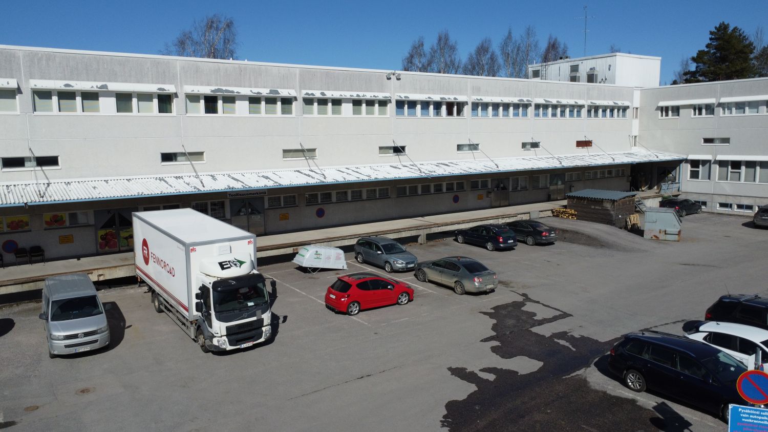 Näkymä teollisuusrakennuksen pihalle, jossa on pysäköitynä kuorma-auto ja useita henkilöautoja.