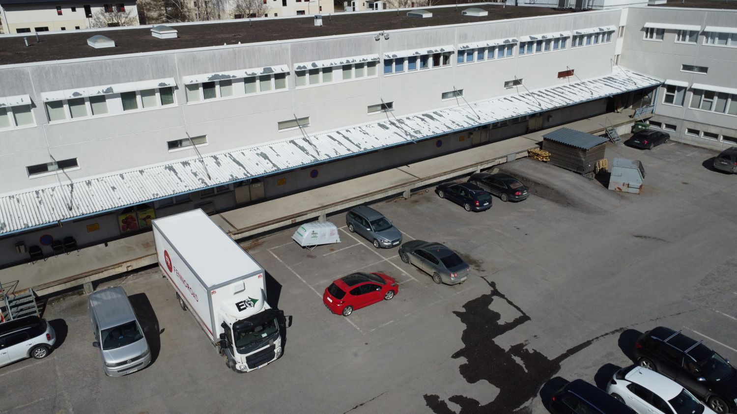 Yläviistosta otettu kuva vaaleasta teollisuusrakennuksesta, jonka pihalla on pysäköityjä autoja j...