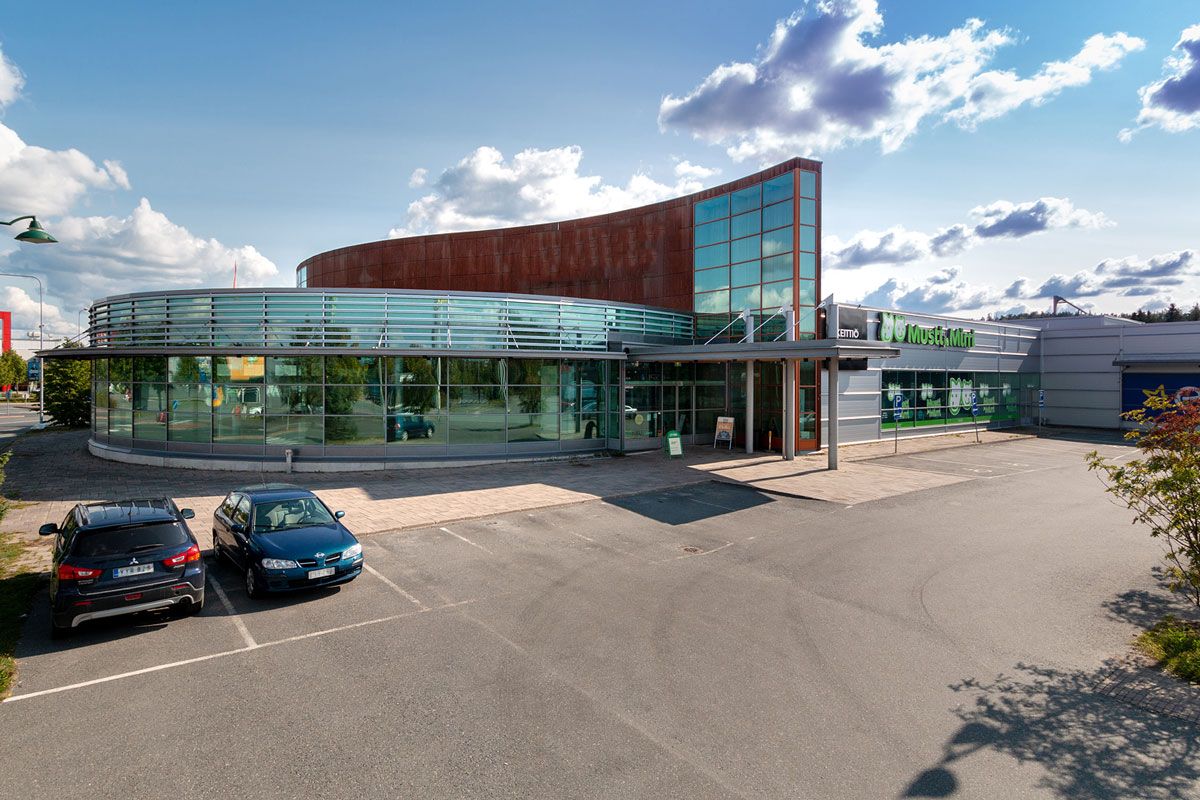 Moderni kaareva liikerakennus ja sen edessä oleva pysäköintialue aurinkoisena päivänä.
