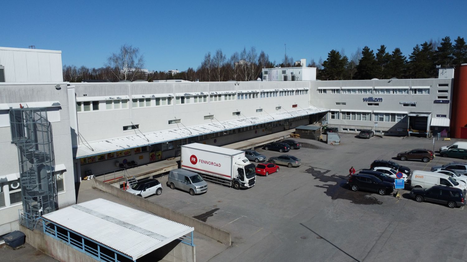 Ilmakuva liikerakennuksesta, jonka pihalla on useita autoja ja lastauslaiturilla oleva rekka.