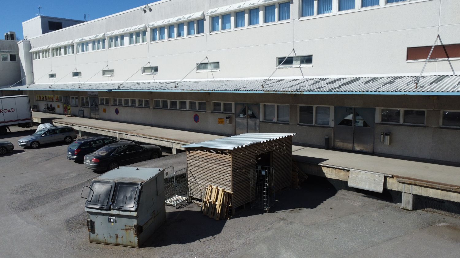 Valkoinen liikerakennus ja sen piha-alue, jossa on lastauslaituri ja pysäköityjä autoja.