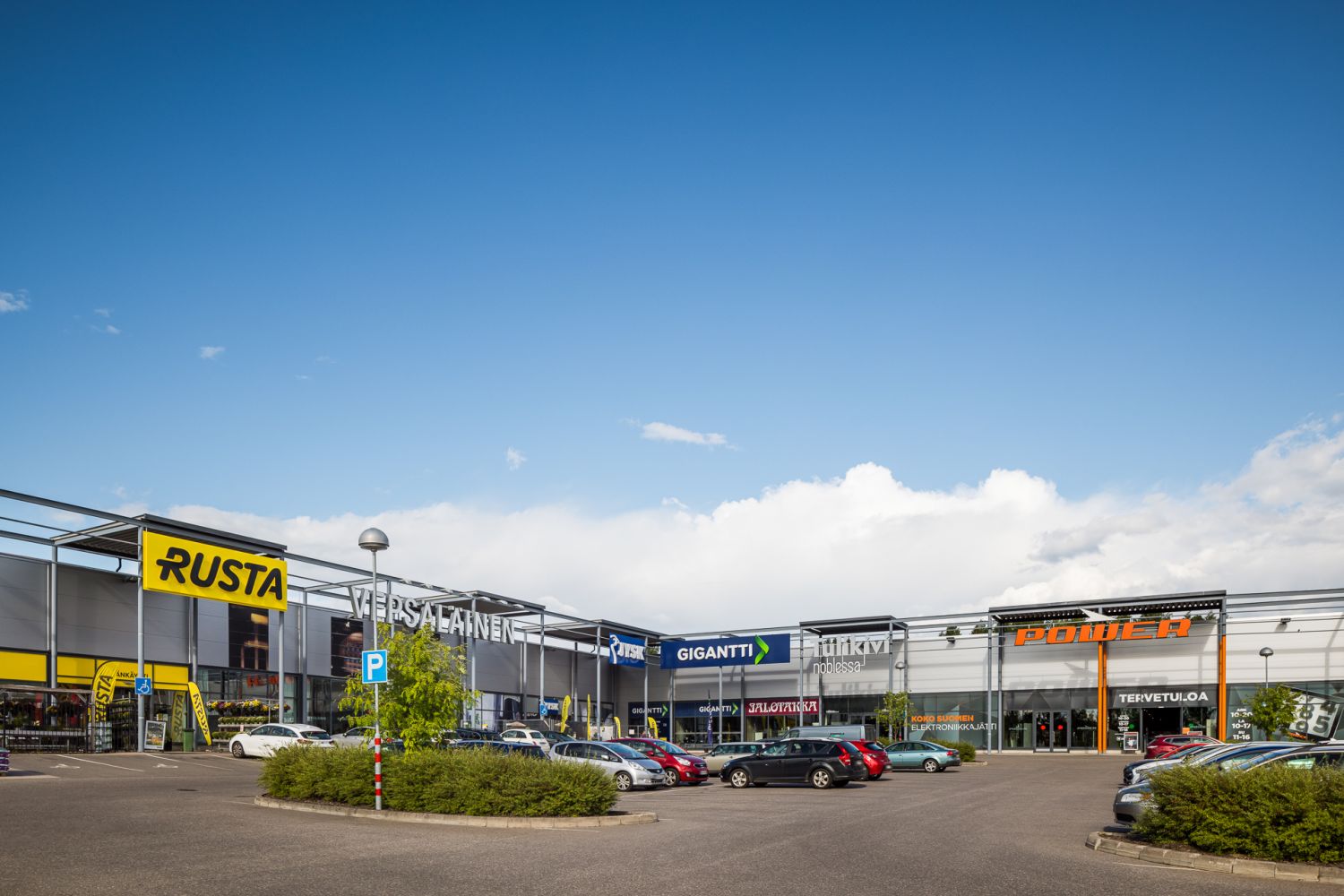 Kauppakeskuksen pysäköintialue ja liikerakennuksia aurinkoisena päivänä.