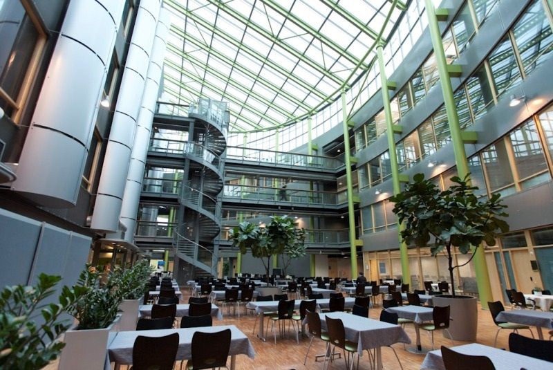 Moderni ja valoisa rakennuksen sisäpiha, jossa on suuri lasikatto, kierreportaat ja ruokailuryhmiä.
