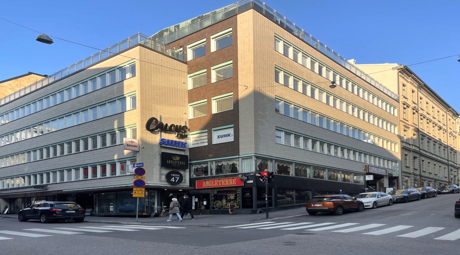 Monikerroksinen rakennus kadunkulmassa aurinkoisena päivänä, kadulla autoja ja jalankulkijoita.