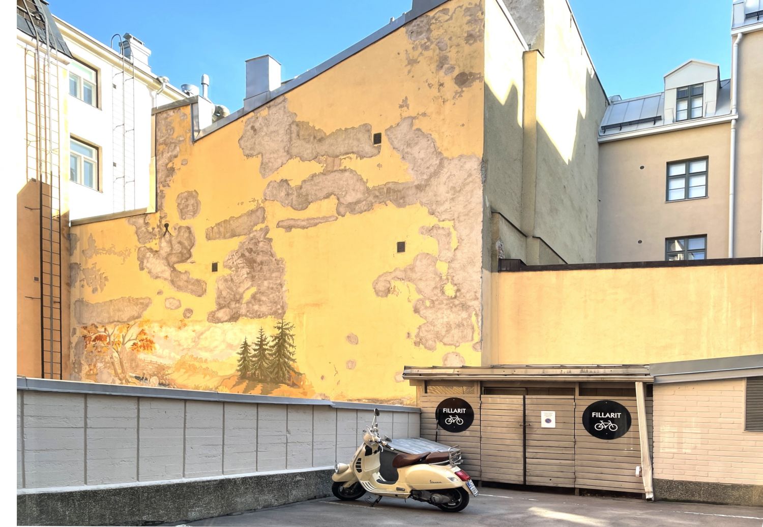 Kermanvärinen skootteri pysäköitynä sisäpihalla suuren keltaisen seinän ja pyörävaraston edessä.