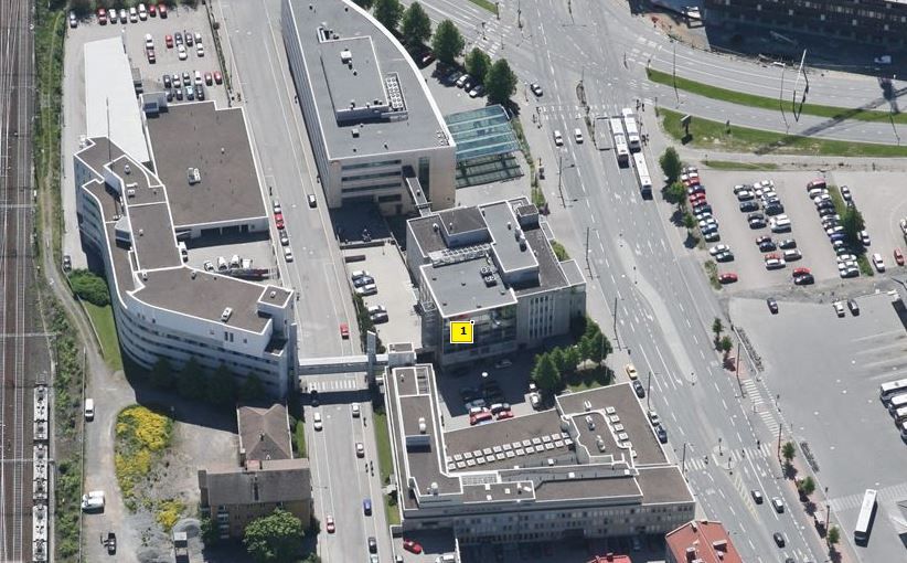 Viisto ilmakuva kaupunkialueesta, jossa on useita rakennuksia, katuja ja pysäköintialueita. Yksi ...
