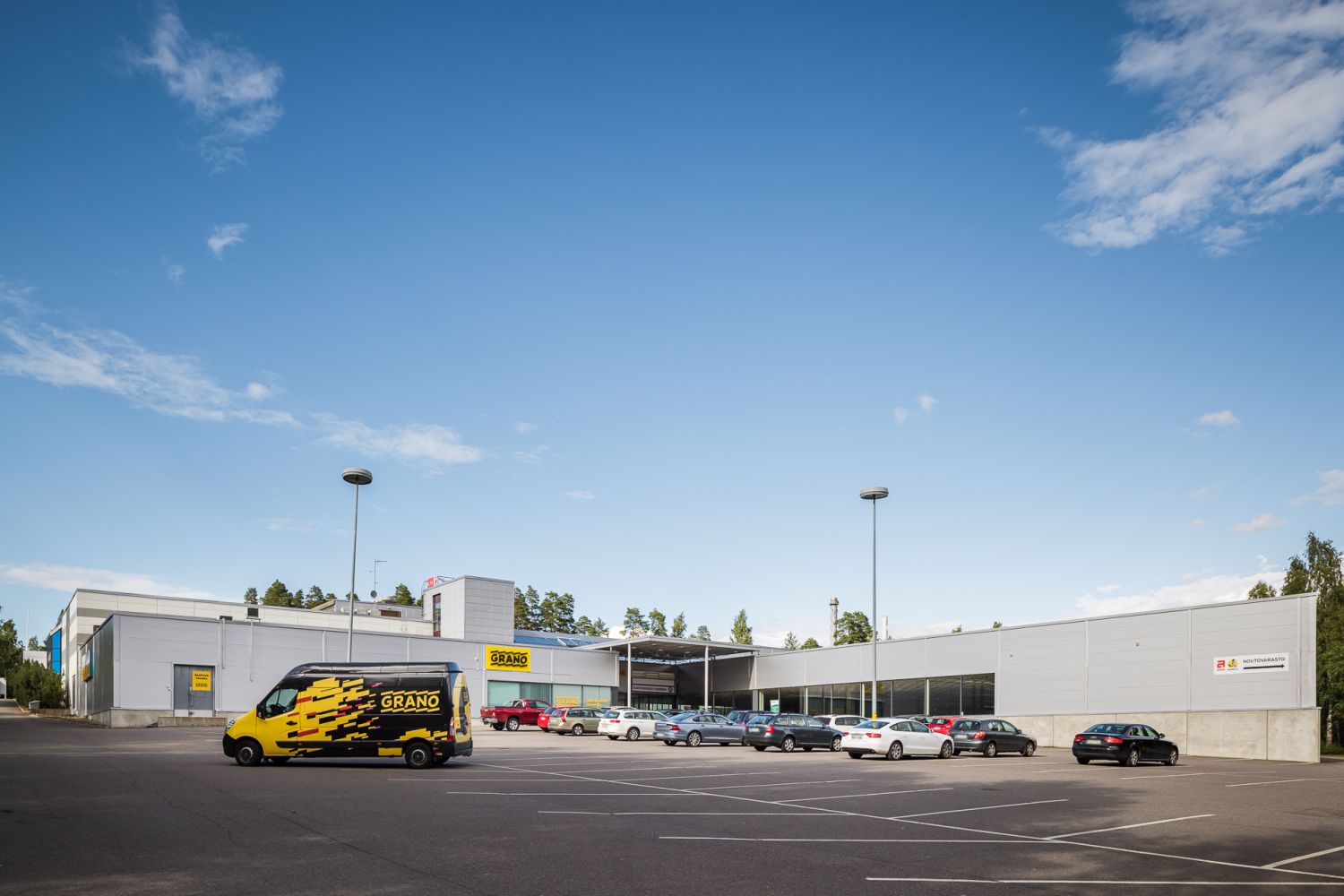 Moderni teollisuusrakennus ja sen parkkipaikka autoineen kirkkaana päivänä.