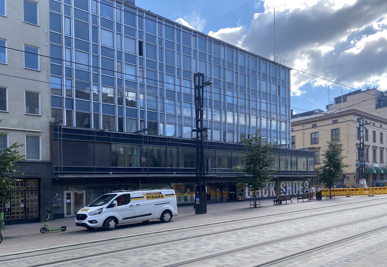 Moderni lasijulkisivuinen rakennus kaupunkikadun varrella, jossa on raitiovaunukiskoja ja valkoin...