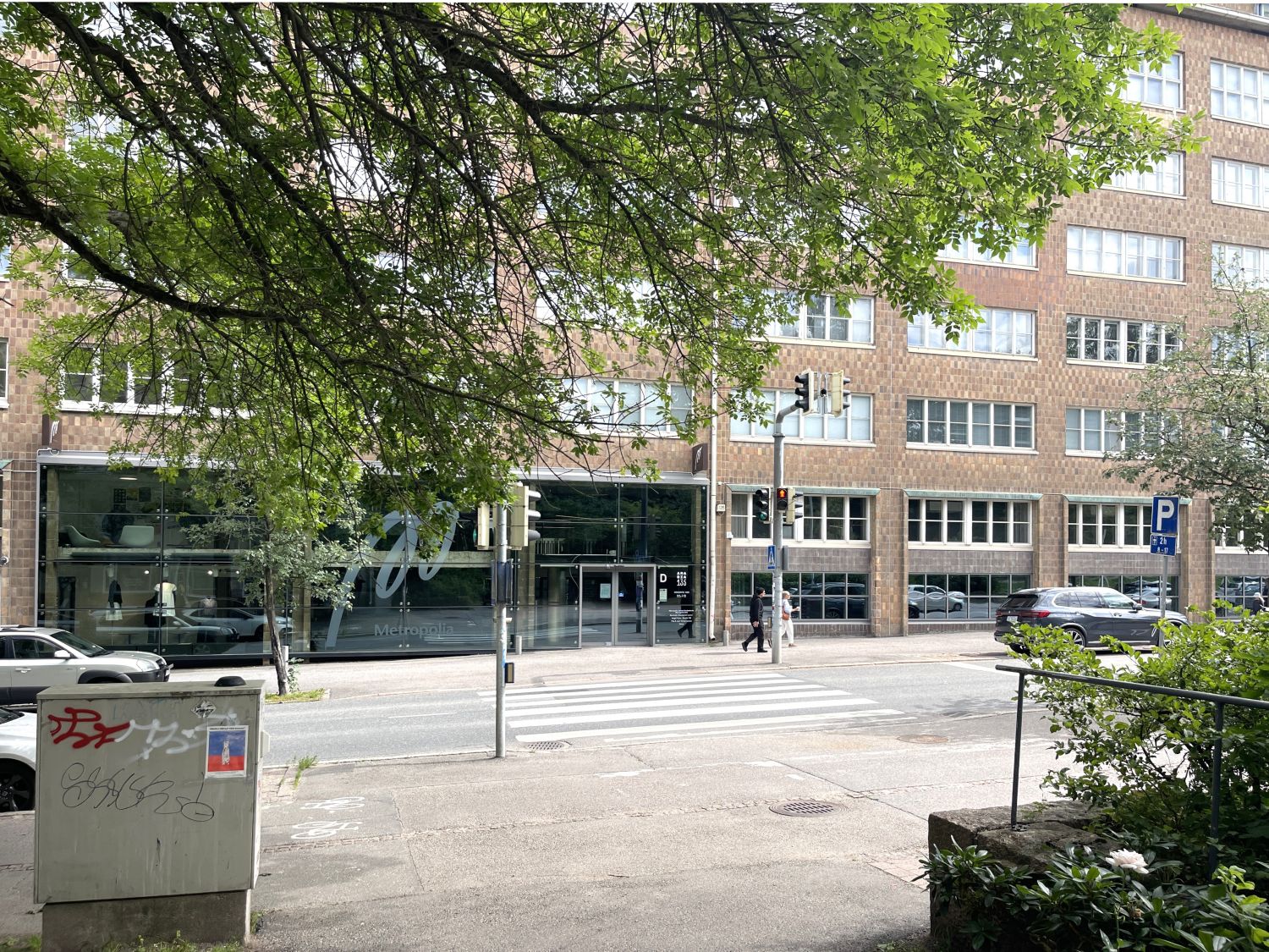 Kaupunkinäkymä, jossa on suuri tiilirakennus, Metropolian sisäänkäynti, suojatie ja puita.