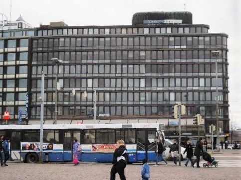 Kaupunkinäkymä, jossa on bussi, jalankulkijoita ja suuri toimistorakennus.