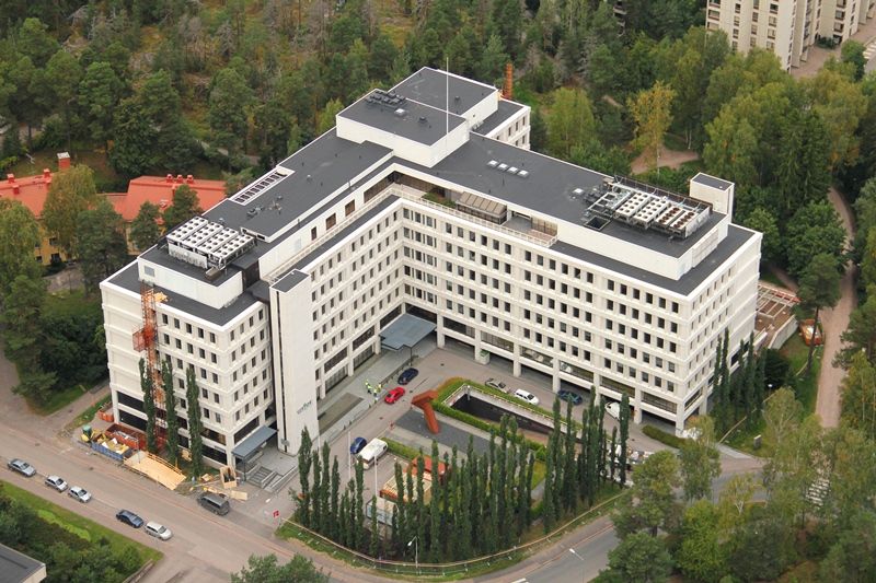 Ilmakuva valkoisesta, monikerroksisesta toimistorakennuksesta, jota ympäröivät vihreät metsäiset ...