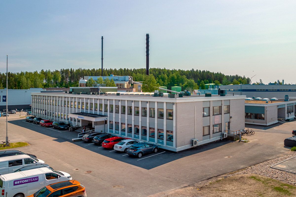 Kaksikerroksinen toimistorakennus ja parkkipaikka teollisuusalueella, taustalla metsää ja savupii...