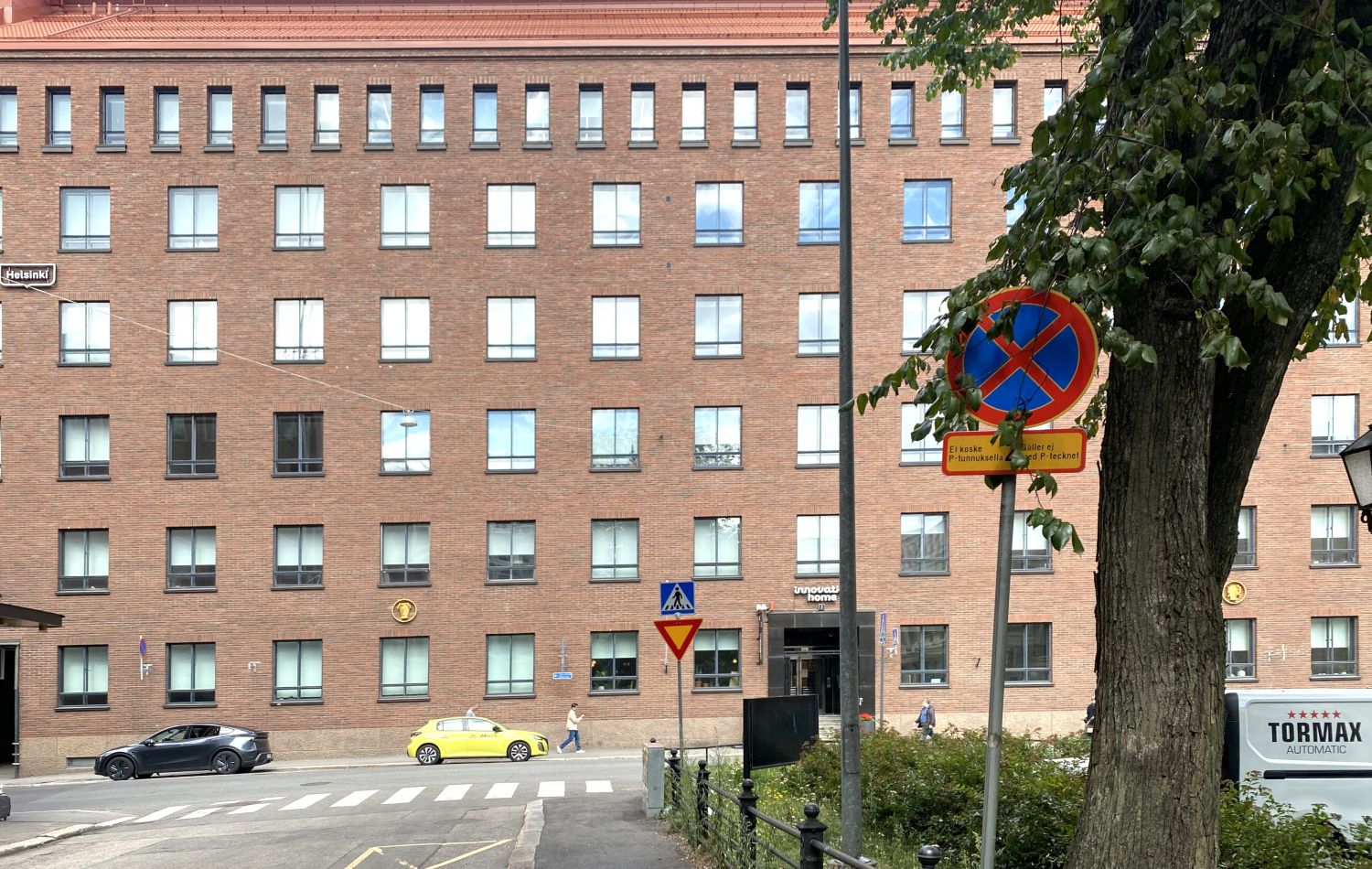 Näkymä katutasolta suureen tiilirakennukseen, jonka edustalla on katu, autoja ja liikennemerkkejä.