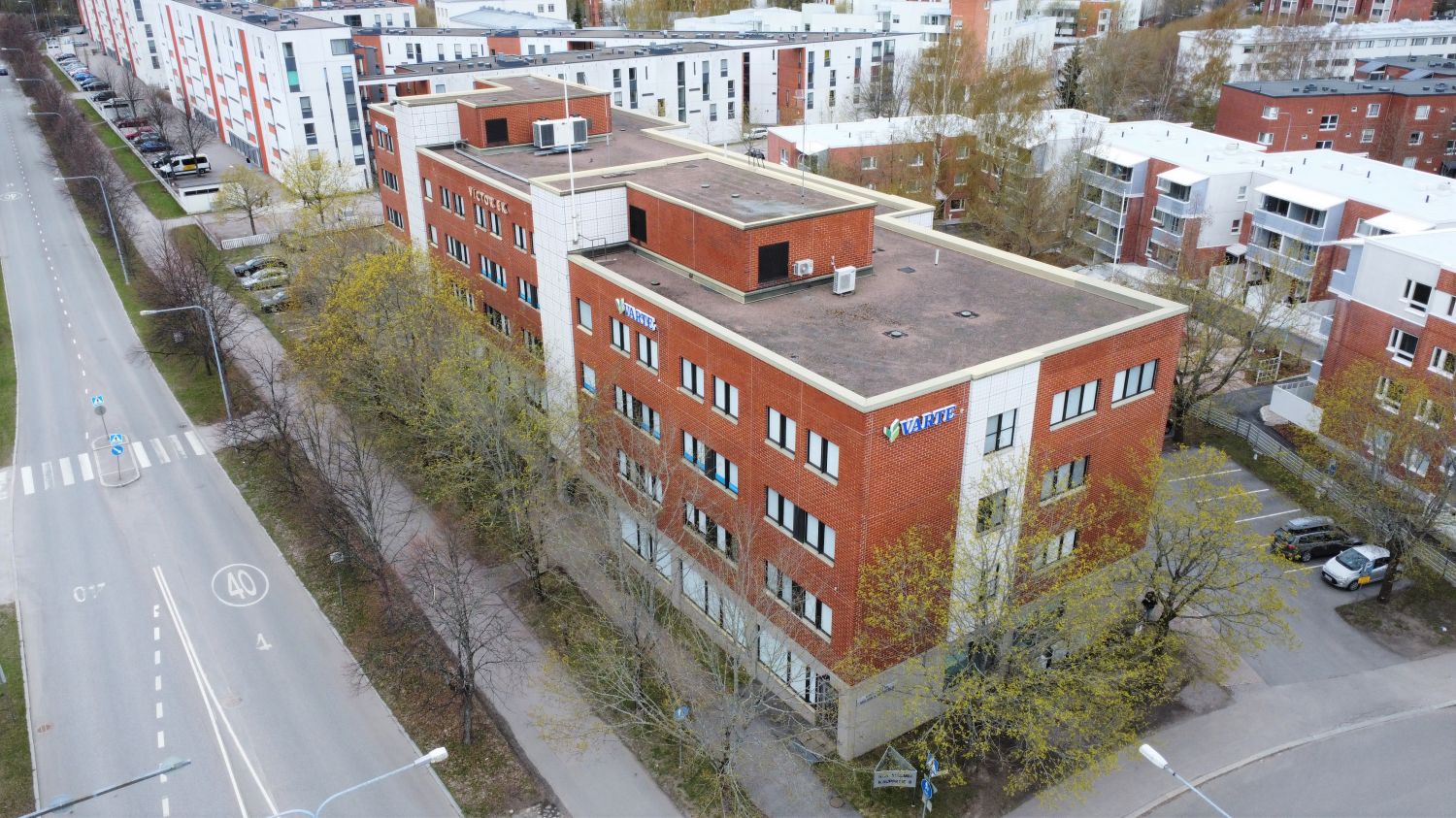 Ilmakuva monikerroksisesta punatiilirakennuksesta ja sitä ympäröivästä kaupunkialueesta.