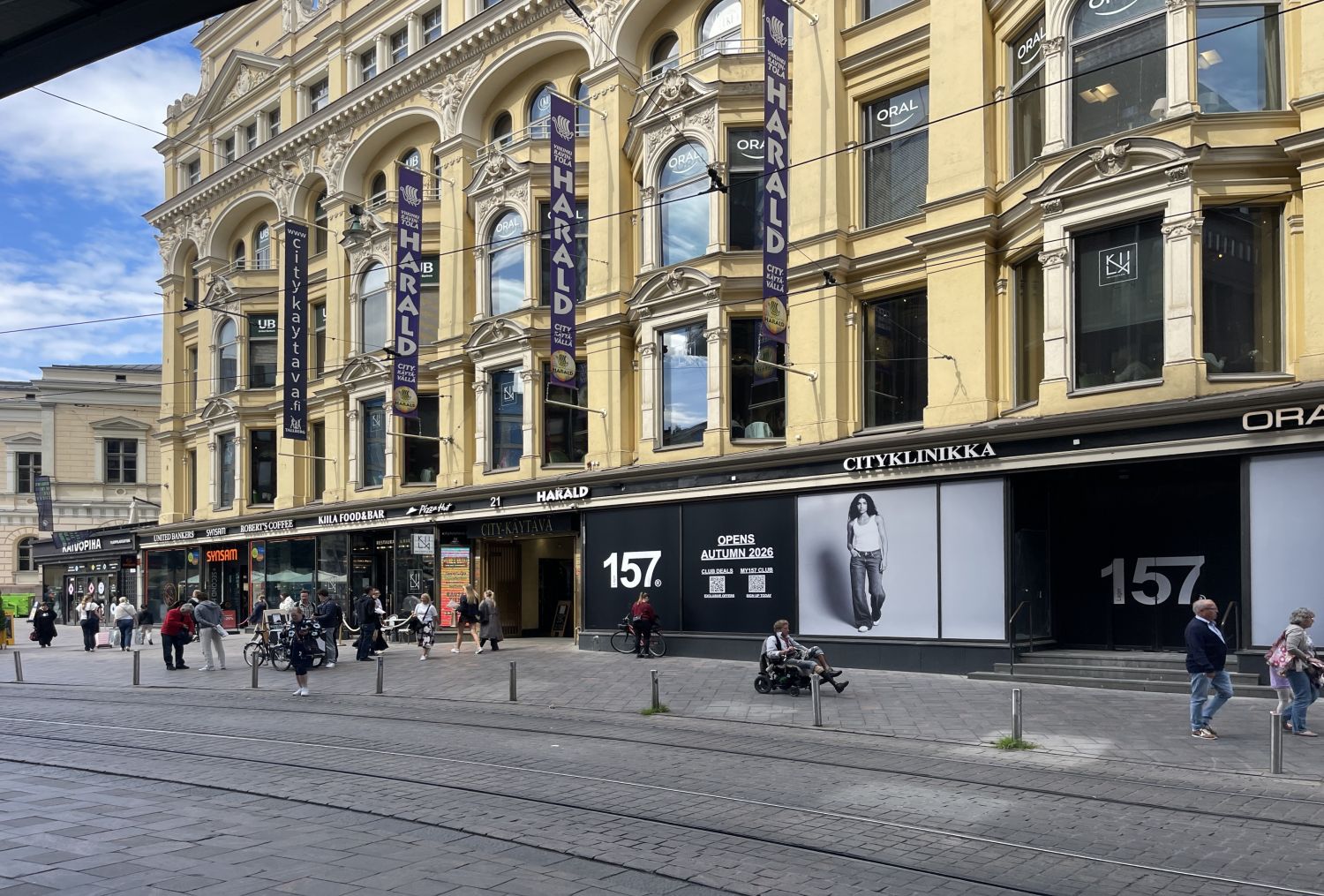 Näkymä vilkkaalta kaupunkikadulta, jossa on koristeellinen keltainen rakennus, liiketiloja ja jal...