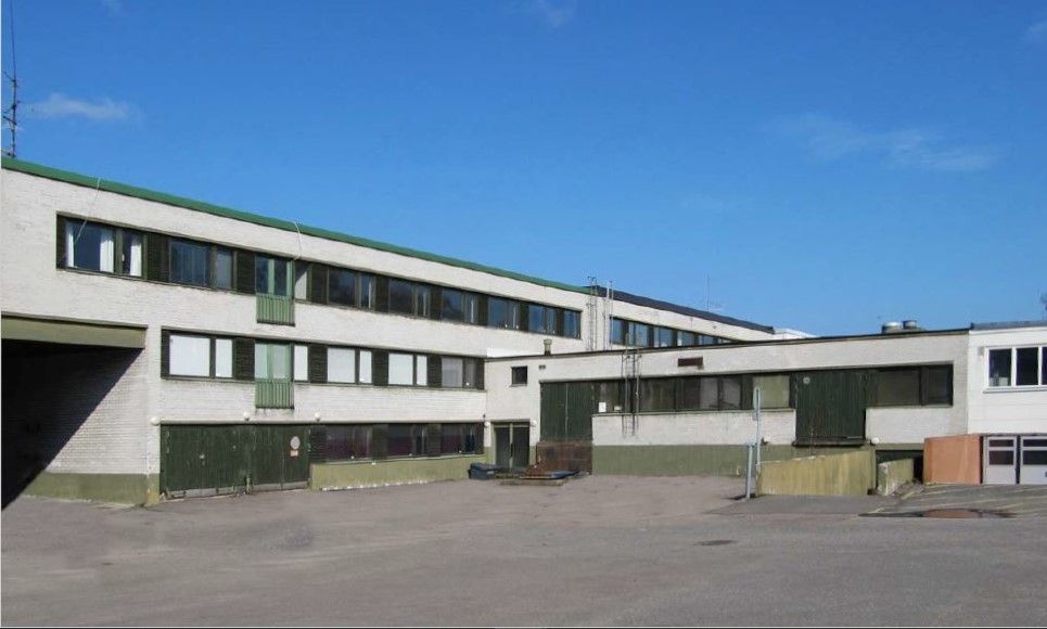 Valkoinen monikerroksinen rakennus, jossa on vihreitä yksityiskohtia, asfaltoidun pihan laidalla.