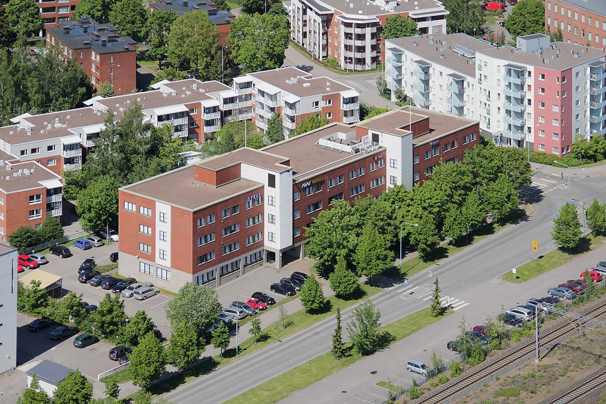 Korkealta otettu ilmakuva kaupunkimaisemasta, jossa on asuin- ja liikerakennuksia, vehreitä puita...