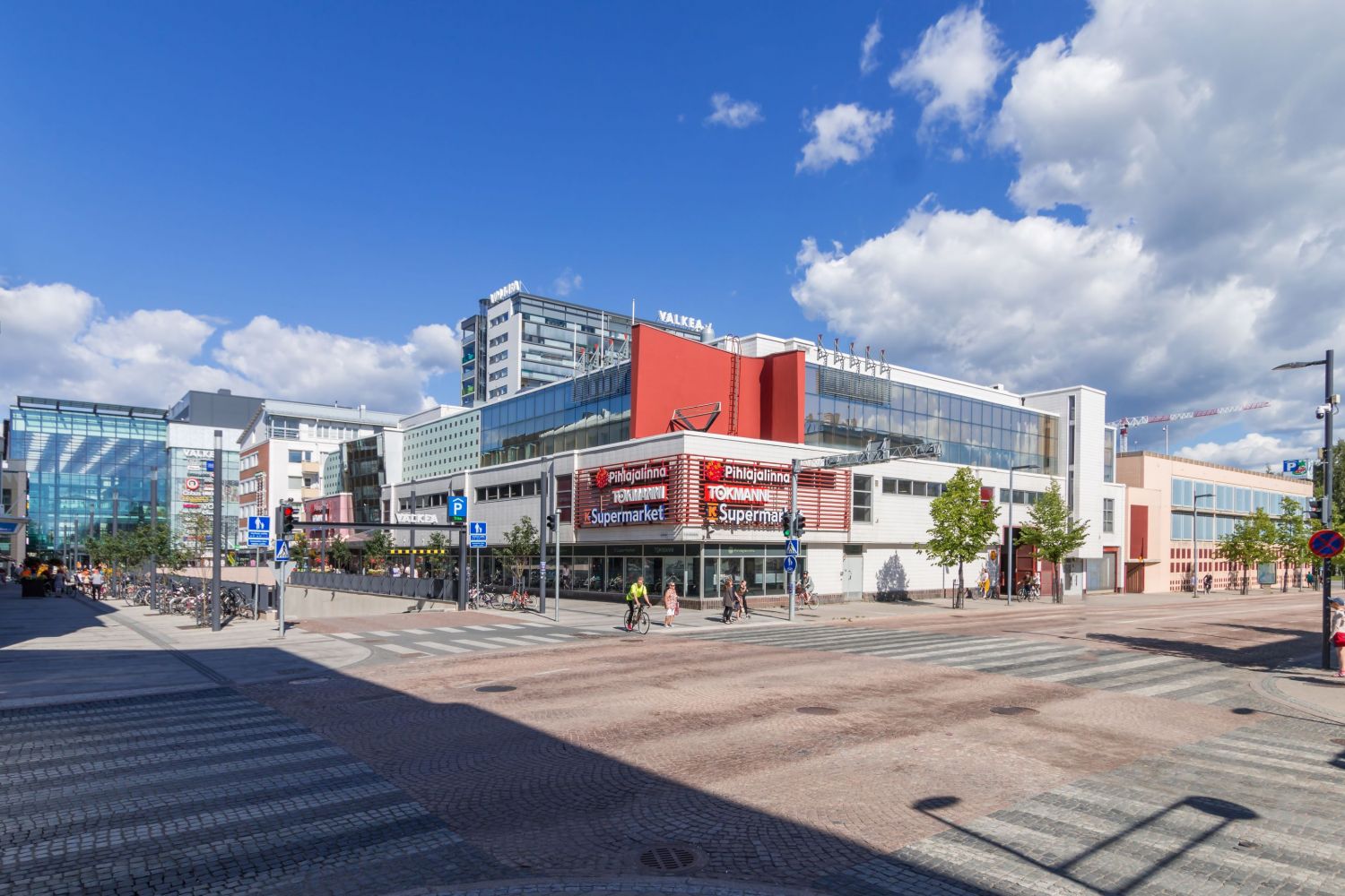 Aurinkoinen kaupunkinäkymä, jossa on moderneja liikerakennuksia, leveä katu ja ihmisiä.