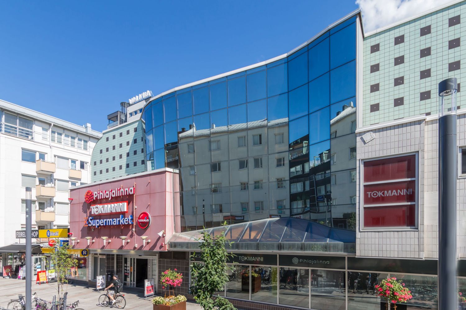 Näkymä kadulta moderniin liikerakennukseen, jonka julkisivussa on Tokmannin, K-Supermarketin ja P...
