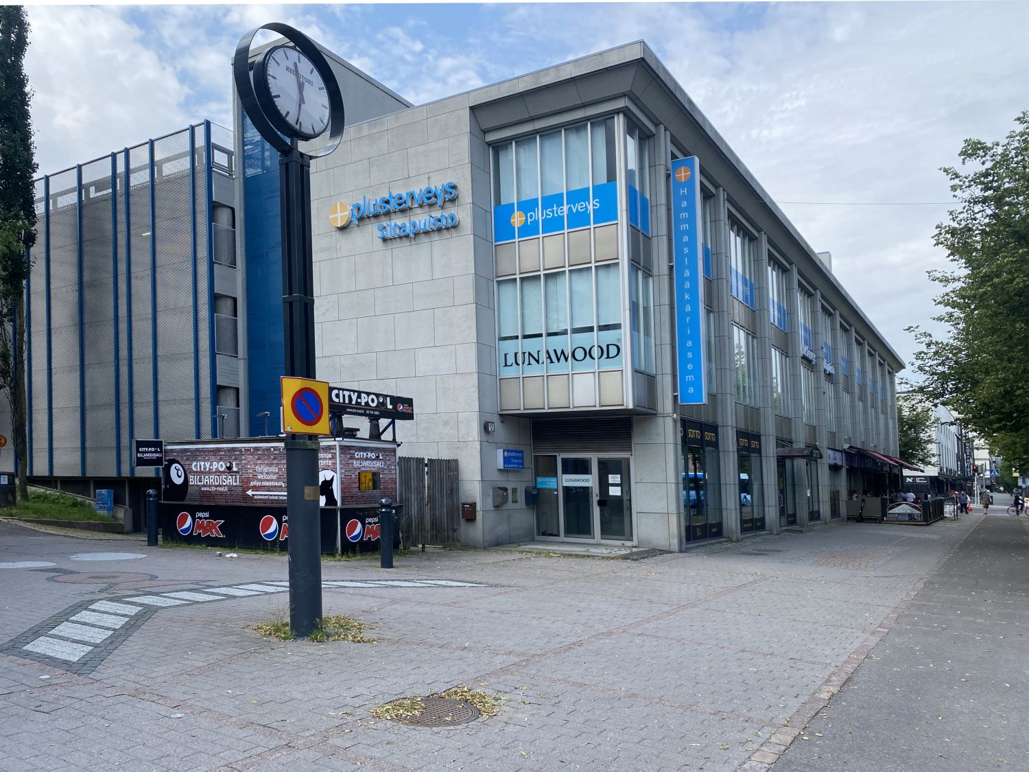 Kaupunkinäkymä liikerakennuksesta, jonka julkisivussa on yritysten kylttejä ja jonka edustalla on...