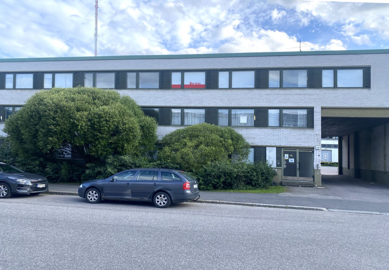 Kaksikerroksinen vaalea rakennus, jonka edessä on pensaita ja kaksi autoa.