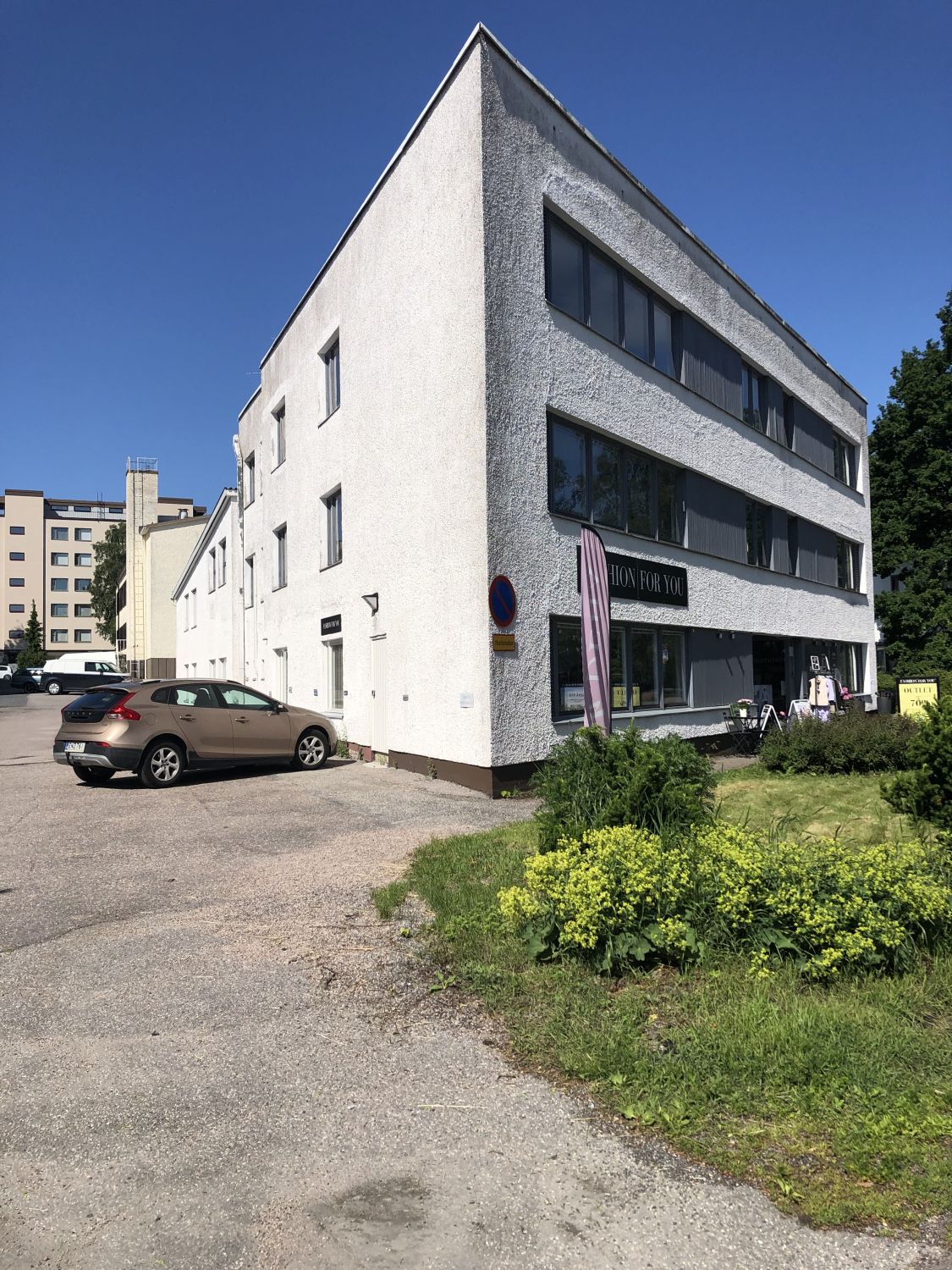 Valkoinen kolmikerroksinen rakennus ja sen edustalle pysäköity auto aurinkoisena päivänä.