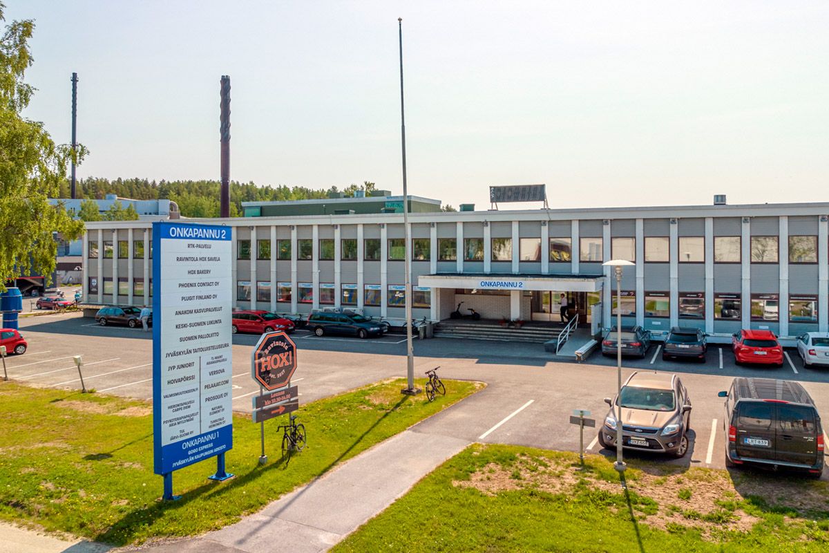 Toimistorakennus Onkapannu 2, sen edessä oleva suuri yrityskyltti ja pysäköintialue autoineen.