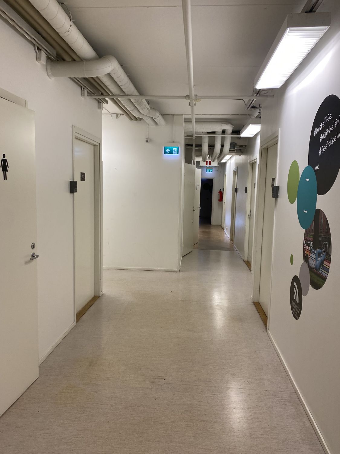 Valkoinen pitkä käytävä, jossa on useita ovia ja katossa kulkevia putkia.