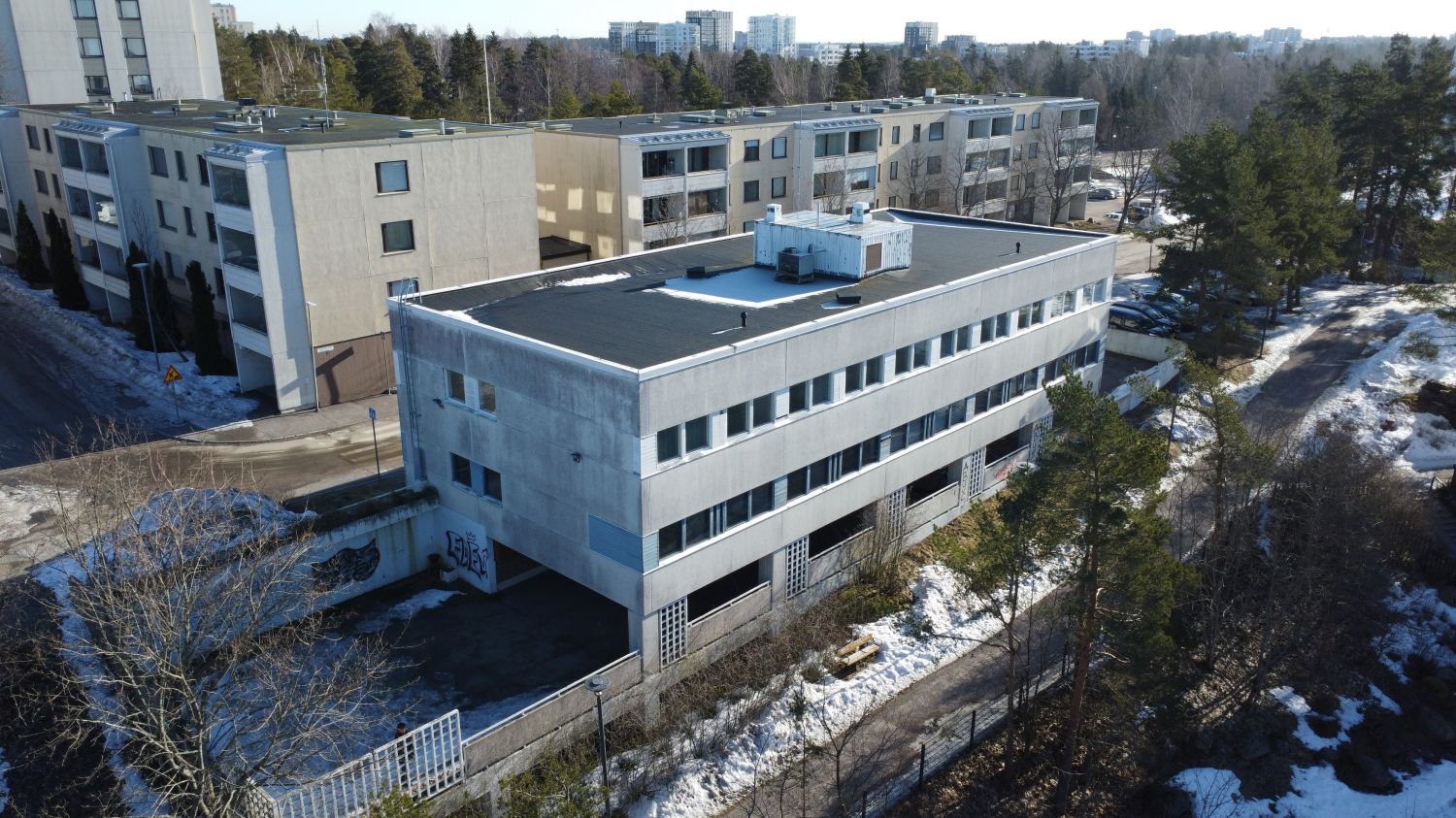 Ilmakuva harmaasta monikerroksisesta rakennuksesta asuinalueella, jossa on vielä hieman lunta maa...