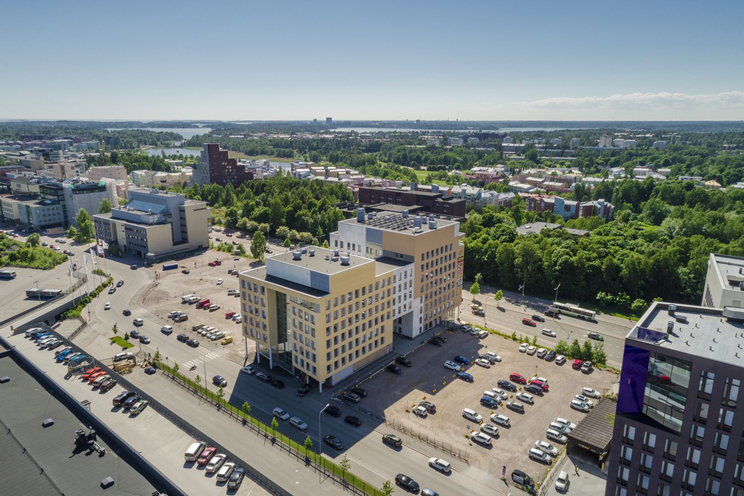 Ilmakuva modernista kaupunkiympäristöstä, jossa on toimistorakennuksia, pysäköintialueita ja vehr...