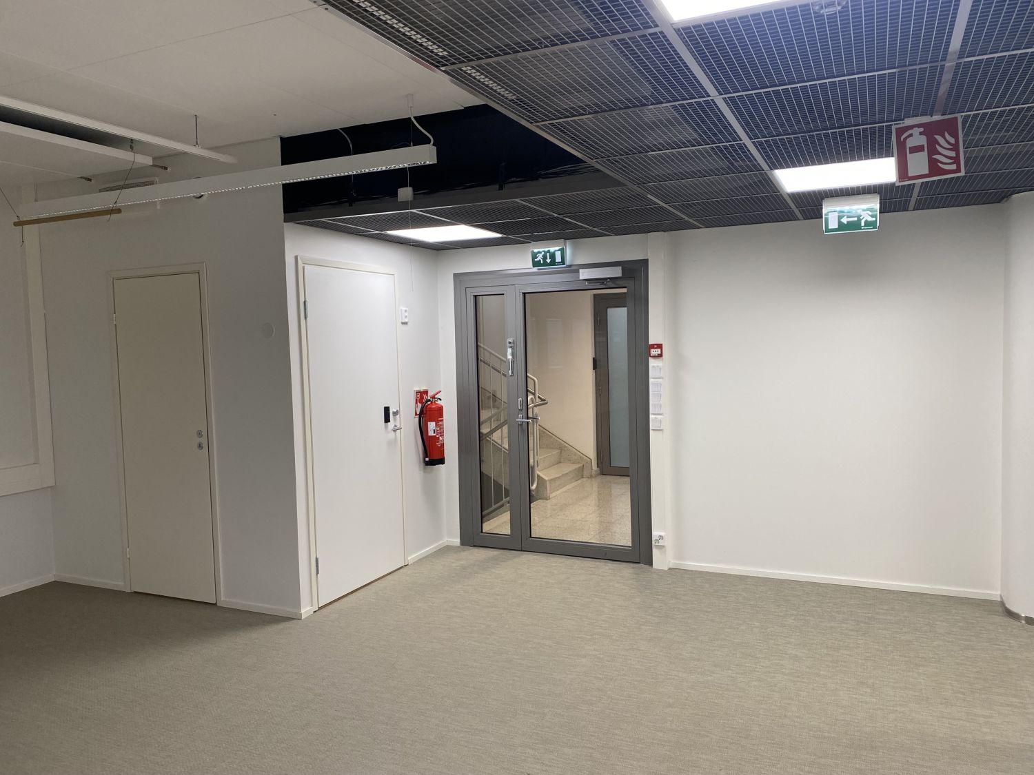 Näkymä rakennuksen sisätiloista, jossa on ovia, lasinen ovi portaikkoon ja poistumistiekylttejä.